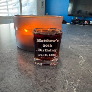 May include: A lit candle in a frosted glass container and a shot glass filled with a dark liquid. The shot glass is engraved with "Matthew's 30th Birthday Dec 21, 2020". The candle and shot glass are on a gray countertop.