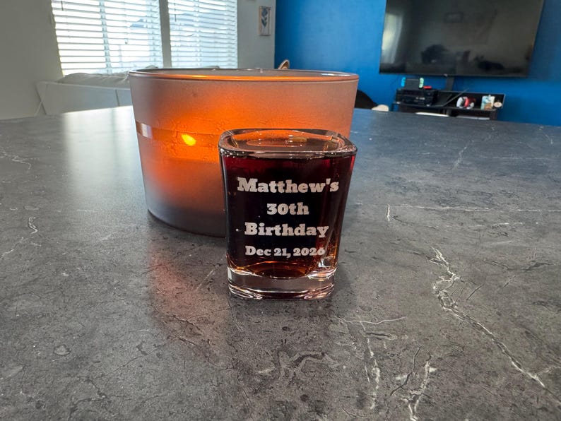 May include: A lit candle in a frosted glass container and a shot glass filled with a dark liquid. The shot glass is engraved with "Matthew's 30th Birthday Dec 21, 2020". The candle and shot glass are on a gray countertop.