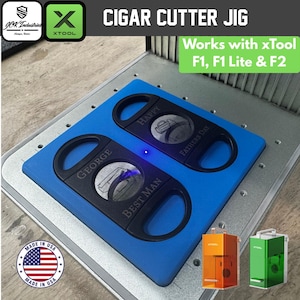 May include: A blue cigar cutter jig featuring two black cigar cutters with engraved text reading GEORGE BEST MAN and HAPPY FATHERS DAY. The jig is compatible with xTool F1, F1 Lite, and F2 machines, and includes a JCK Industries logo and a USA flag emblem.