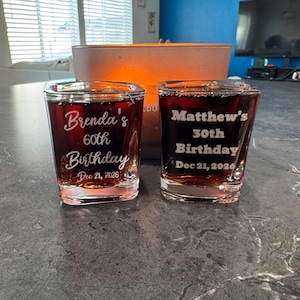 May include: Two clear shot glasses filled with a dark liquid. One glass is inscribed with "Brenda's 60th Birthday Dec 21, 2026." The other glass reads "Matthew's 30th Birthday Dec 21, 2026."