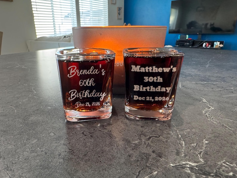 May include: Two clear shot glasses filled with a dark liquid. One glass is inscribed with "Brenda's 60th Birthday Dec 21, 2026." The other glass reads "Matthew's 30th Birthday Dec 21, 2026."