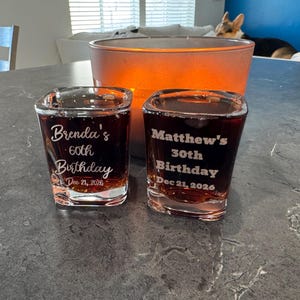 May include: Two square shot glasses filled with a dark liquid, each engraved with a birthday message. One reads "Brenda's 60th Birthday Dec 21, 2026", the other "Matthew's 30th Birthday Dec 21, 2026". A large orange candle holder is in the background.