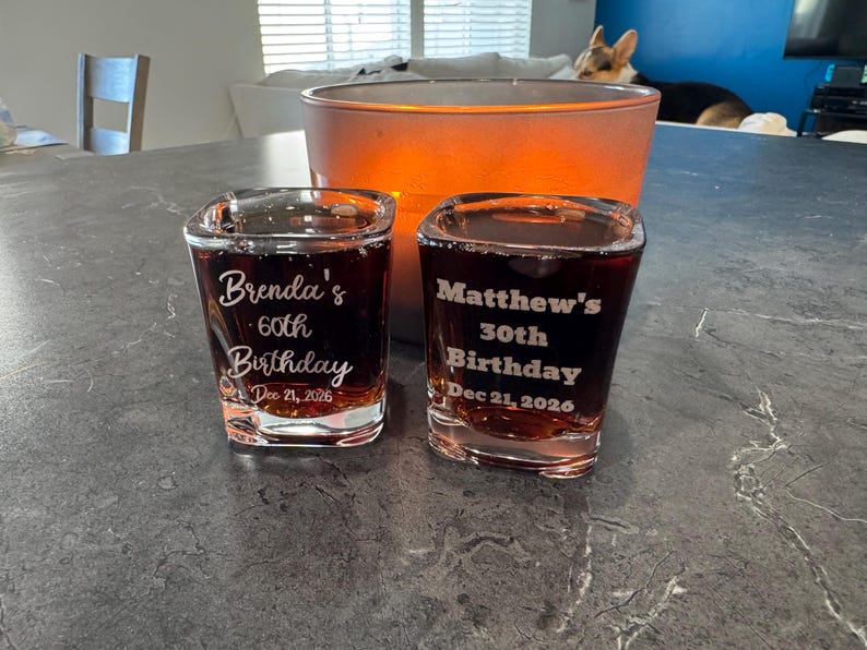 May include: Two square shot glasses filled with a dark liquid, each engraved with a birthday message. One reads "Brenda's 60th Birthday Dec 21, 2026", the other "Matthew's 30th Birthday Dec 21, 2026". A large orange candle holder is in the background.