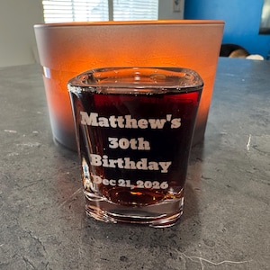 May include: A clear shot glass filled with a dark liquid, featuring the etched text "Matthew's 30th Birthday Dec 21, 2026." The glass is in front of an orange and brown container. The shot glass is on a gray surface.