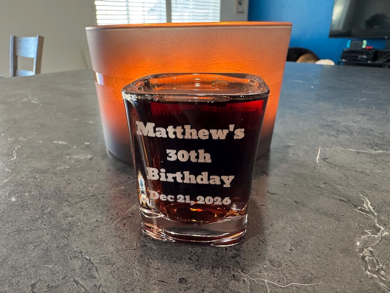 May include: A clear shot glass filled with a dark liquid, featuring the etched text "Matthew's 30th Birthday Dec 21, 2026." The glass is in front of an orange and brown container. The shot glass is on a gray surface.