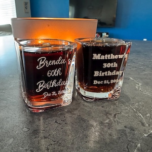 May include: Two clear shot glasses filled with a dark liquid. One glass reads "Brenda's 60th Birthday Dec 21, 2026" and the other reads "Matthew's 30th Birthday Dec 21, 2026".