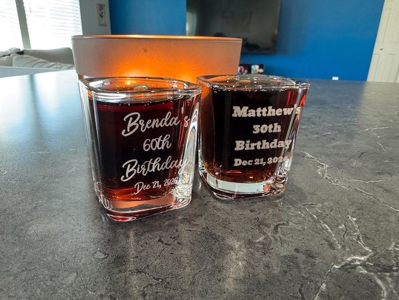 May include: Two clear shot glasses filled with a dark liquid. One glass reads "Brenda's 60th Birthday Dec 21, 2026" and the other reads "Matthew's 30th Birthday Dec 21, 2026".