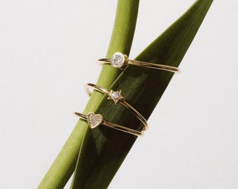 May include: Three delicate gold rings, each with a unique design: a round diamond, a star, and a heart. The rings are displayed on a vibrant green leaf against a white background, showcasing their elegant simplicity.