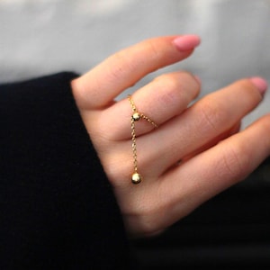 Könnte beinhalten: Ein zarter goldener Ring mit einer Kette und zwei kleinen Kugeln. Der Ring wird an einem Finger getragen, wobei die Kette und die Kugeln herunterhängen. Ein schlichtes, elegantes Schmuckstück.