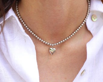May include: A silver beaded necklace with a heart pendant. The necklace is composed of small, round beads and features a heart-shaped charm at the center. The necklace is worn over a white collared shirt.