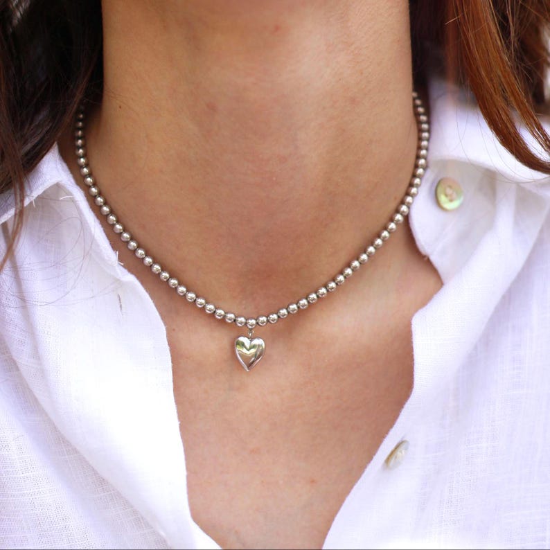 Op de afbeelding: Zilveren ketting met een hartvormige hanger. De ketting bestaat uit kleine, ronde kralen en heeft een hartvormige bedel. De ketting wordt gedragen over een wit overhemd met kraag.