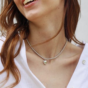 Op de afbeelding: Een zilveren kralenketting met een kleine hartvormige hanger. De ketting wordt gedragen over een wit overhemd. De hartvormige hanger is gecentreerd en hangt onder het sleutelbeen. De kralen zijn klein en gelijkmatig verdeeld.