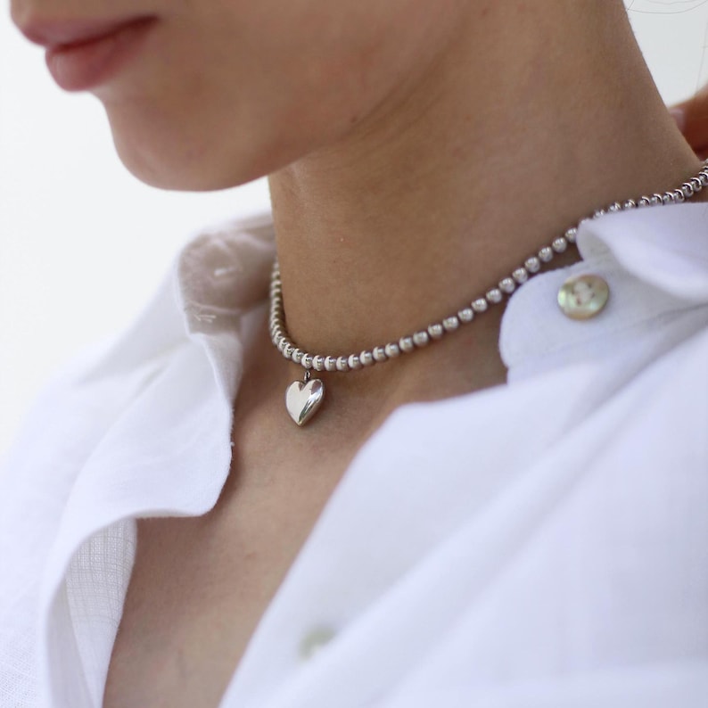 Op de afbeelding: Een zilveren ketting met een kralenketting en een hartvormige hanger. De ketting wordt gedragen over een wit overhemd. De hartvormige hanger is glad en reflecterend.