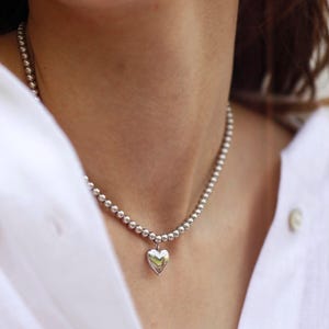 Op de afbeelding: Zilveren ketting met een hartvormige hanger, bestaande uit kleine, ronde kralen. De hartvormige bedel is gepolijst. De ketting wordt gedragen op een wit overhemd.