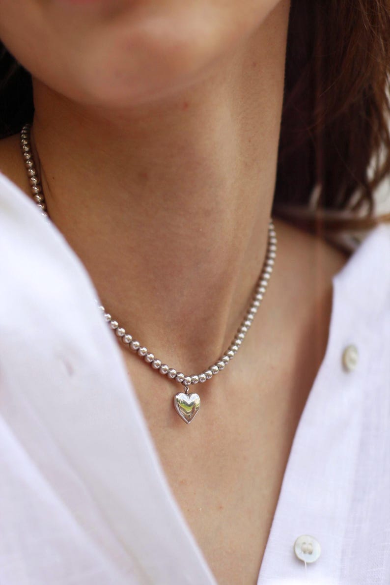 Op de afbeelding: Zilveren ketting met een hartvormige hanger, bestaande uit kleine, ronde kralen. De hartvormige bedel is gepolijst. De ketting wordt gedragen op een wit overhemd.