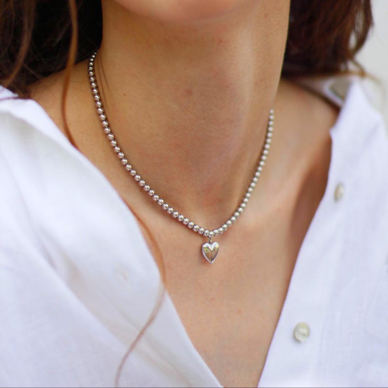 Op de afbeelding: Een zilveren kralenketting met een hartvormige hanger. De ketting bestaat uit kleine, ronde kralen. De hartvormige hanger is glad en gepolijst. De achtergrond is een wit overhemd.