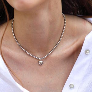 Op de afbeelding: Een zilveren kralenketting met een hartvormige hanger. De ketting bestaat uit kleine, ronde kralen en hangt om de nek. De hartvormige hanger is gecentreerd en hangt onder de halslijn. De ketting wordt gedragen over een wit overhemd.