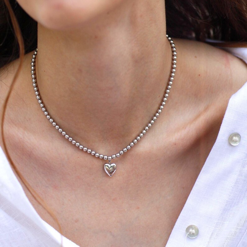 Op de afbeelding: Een zilveren kralenketting met een hartvormige hanger. De ketting bestaat uit kleine, ronde kralen en hangt om de nek. De hartvormige hanger is gecentreerd en hangt onder de halslijn. De ketting wordt gedragen over een wit overhemd.