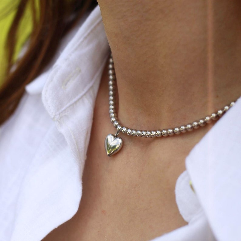 Op de afbeelding: Een zilveren kralenketting met een hartvormige hanger. De ketting wordt gedragen tegen een wit overhemd. De hartvormige hanger is glad en reflecterend. De kralen zijn klein en gelijkmatig verdeeld.