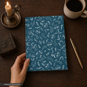 May include: A teal notebook with a white mushroom and paisley pattern. The notebook is held by a hand on a dark wooden surface. A lit candle, a cup of coffee, and a gold pen are also on the table.