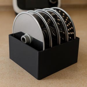 Könnte beinhalten: Ein schwarzer rechteckiger Behälter mit mehreren Edelstahl-Küchenmaschinenklingen. Die Klingen haben unterschiedliche Designs, darunter durchgehende, geschlitzte und perforierte Oberflächen. Der Behälter und die Klingen stehen auf einer hellbraunen Oberfläche.