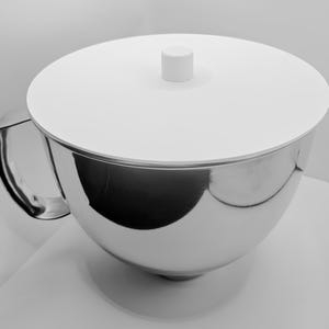 Könnte beinhalten: Eine Edelstahlschüssel mit weißem Deckel und Griff. Die Schüssel hat eine abgerundete Form und eine glänzende, reflektierende Oberfläche. Der Deckel hat einen kleinen, zylindrischen Knopf in der Mitte. Der Griff ist an der Seite der Schüssel befestigt.