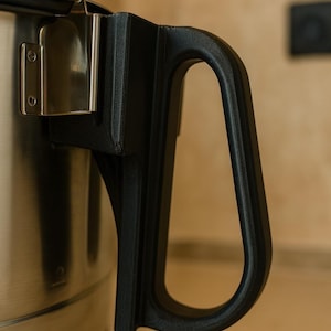 Op de afbeelding: Close-up van een roestvrijstalen pot met een zwarte handgreep. De handgreep heeft een stevig, gebogen ontwerp en is met een metalen beugel aan de pot bevestigd. Het oppervlak van de pot is glad en reflecterend.