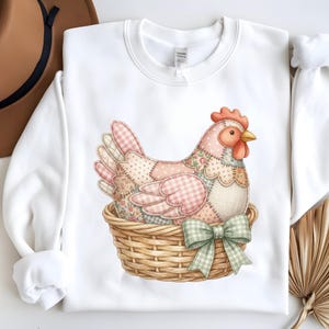 May include: White sweatshirt with a detailed illustration of a patchwork hen in a woven basket. The hen has pink, floral, and gingham patterned feathers, a red comb, and a yellow beak. A green and white checkered bow adorns the basket.