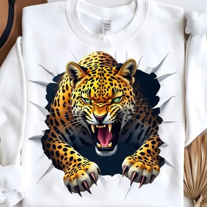 Puede incluir: Sudadera blanca con una ilustración detallada de un leopardo rompiendo una superficie rasgada. El leopardo tiene una expresión feroz, ojos verdes y la boca abierta, mostrando sus dientes. El diseño es audaz y llamativo.