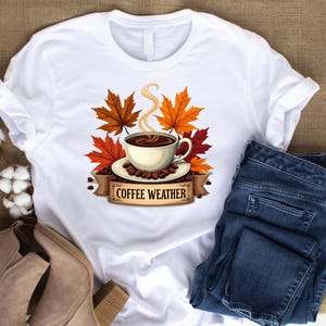 May include: White t-shirt with a graphic of a coffee cup with steam, surrounded by autumn leaves and coffee beans. The text "Coffee Weather" is printed below the cup.