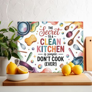 May include: A rectangular kitchen sign with the text "The Secret to a Clean Kitchen is simple: Don't Cook (Ever!)". The sign has a colorful design with kitchen utensils and food illustrations. Lemons in a white bowl and on a wooden cutting board are in the foreground.