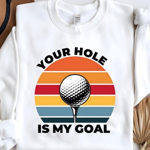 Könnte beinhalten: Weißes Sweatshirt mit einer Grafik eines Golfballs auf einem Tee, vor einem farbenfrohen Sonnenuntergangsdesign. Der Text "YOUR HOLE IS MY GOAL" ist oberhalb und unterhalb des Golfballbildes aufgedruckt.