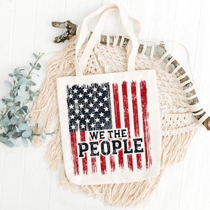 Può includere: Una borsa tote in tela bianca con un design di bandiera americana usurato e il testo "WE THE PEOPLE" in nero. La borsa è appoggiata su una superficie bianca con un pezzo di legno alla deriva e un arazzo macramé.