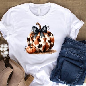 May include: White t-shirt with a brown and black patterned pumpkin with a black bow. The pumpkin is surrounded by small brown leaves. A smaller orange pumpkin is in the foreground.