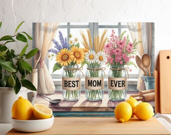 May include: A canvas print featuring three mason jars filled with flowers: sunflowers, daisies, and pink blooms. Each jar has a label that reads "BEST MOM EVER." The background shows a window with curtains and kitchen utensils.