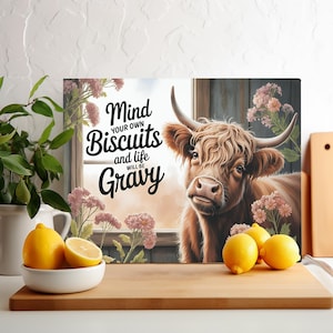 Può includere: Una mucca Highland marrone e bianca con pelo lungo e arruffato guarda la telecamera. La mucca è davanti a una finestra con un disegno floreale e il testo "Mind your own Biscuits and life will be Gravy".