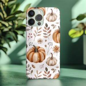 May include: A white phone case featuring a watercolor pattern of pumpkins, leaves, and flowers in shades of orange, brown, and beige. The case has a glossy finish and a camera cutout.