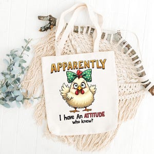 Può includere: Borsa tote in tela bianca con un pollo dei cartoni animati che indossa un fiocco verde. Il testo sulla borsa recita "Apparentemente ho un atteggiamento, chi lo sapeva?"