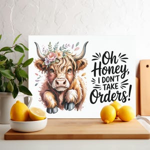 May include: A watercolor print of a Highland cow with a floral crown. The cow has brown fur and large horns. Text on the print reads, "Oh Honey, I don't take orders!" Lemons in a white bowl and on a wooden cutting board are in the foreground.