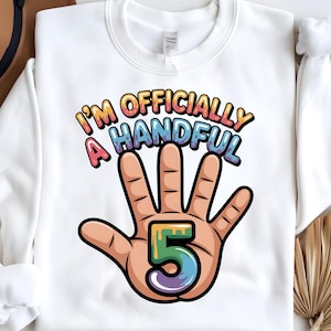 May include: White sweatshirt featuring the phrase "I'M OFFICIALLY A HANDFUL" in colourful bubble letters. A cartoon hand with the number "5" in the centre completes the design. A fun birthday shirt for a five-year-old.