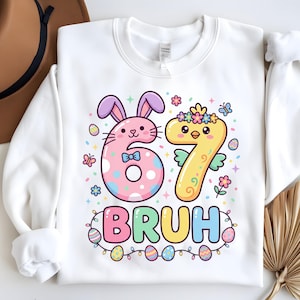 Pode incluir: Camisola branca com um gráfico colorido com tema de Páscoa. O design apresenta os números "67" com um coelho e um pintinho, a palavra "BRUH" e ovos de Páscoa. O coelho é rosa com orelhas roxas e o pintinho é amarelo.