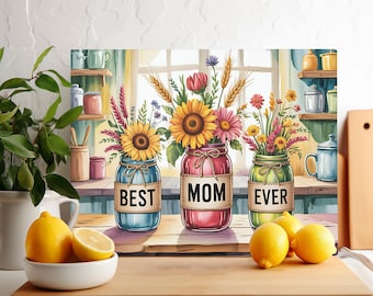 May include: A decorative kitchen cutting board featuring three mason jars filled with colorful flowers. The jars are labeled "BEST", "MOM", and "EVER". The background includes a window, shelves, and lemons.