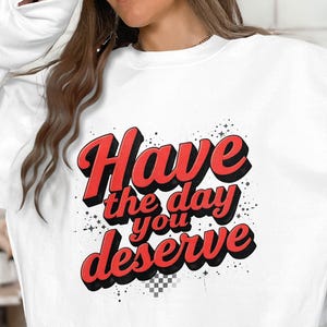Może przedstawiać: Biała bluza z napisem "Have the day you deserve" w kolorze czerwonym, obrysowanym na czarno, w stylu retro. Projekt zawiera małe akcenty w kształcie gwiazd i wzór w szachownicę.