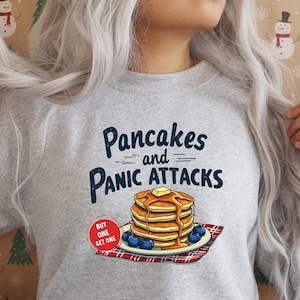 May include: A gray sweatshirt with the text "Pancakes and Panic Attacks" in black and white. The text is in a retro font. There is a cartoon image of a stack of pancakes with blueberries and syrup on a red and white checkered plate. The text "Buy One Get One" is in a red circle.
