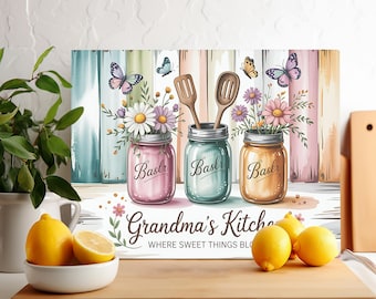 May include: A decorative kitchen sign featuring three mason jars filled with flowers and wooden utensils. The jars are in shades of pink, blue, and orange, with the word "Baslr" on each. The sign reads "Grandma's Kitchen" with the phrase "WHERE SWEET THINGS BLOOM".