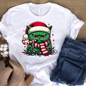 May include: White t-shirt featuring a green cat wearing a red and white Santa hat and scarf. The cat is holding a candy cane and is surrounded by Christmas lights.