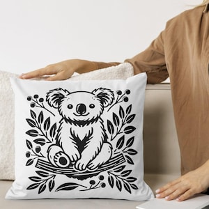 May include: A white throw pillow with a black and white illustration of a koala sitting on a branch with leaves. The koala is surrounded by leaves and berries.