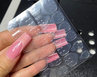 May include: A sheet of clear adhesive tabs for attaching fake nails. The tabs are in various shapes and sizes, and there are several pink fake nails on the sheet.