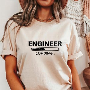 May include: Beige t-shirt with the text "ENGINEER LOADING..." in black. The word "ENGINEER" is above a loading bar graphic. The shirt has short sleeves and a relaxed fit.