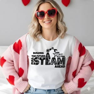 May include: A white t-shirt with the words "FULL STEAM AHEAD" in black, along with the text "BUILDING THE FUTURE" and "one STAR at a time". The person is wearing a pink cardigan with red hearts and red heart-shaped sunglasses.